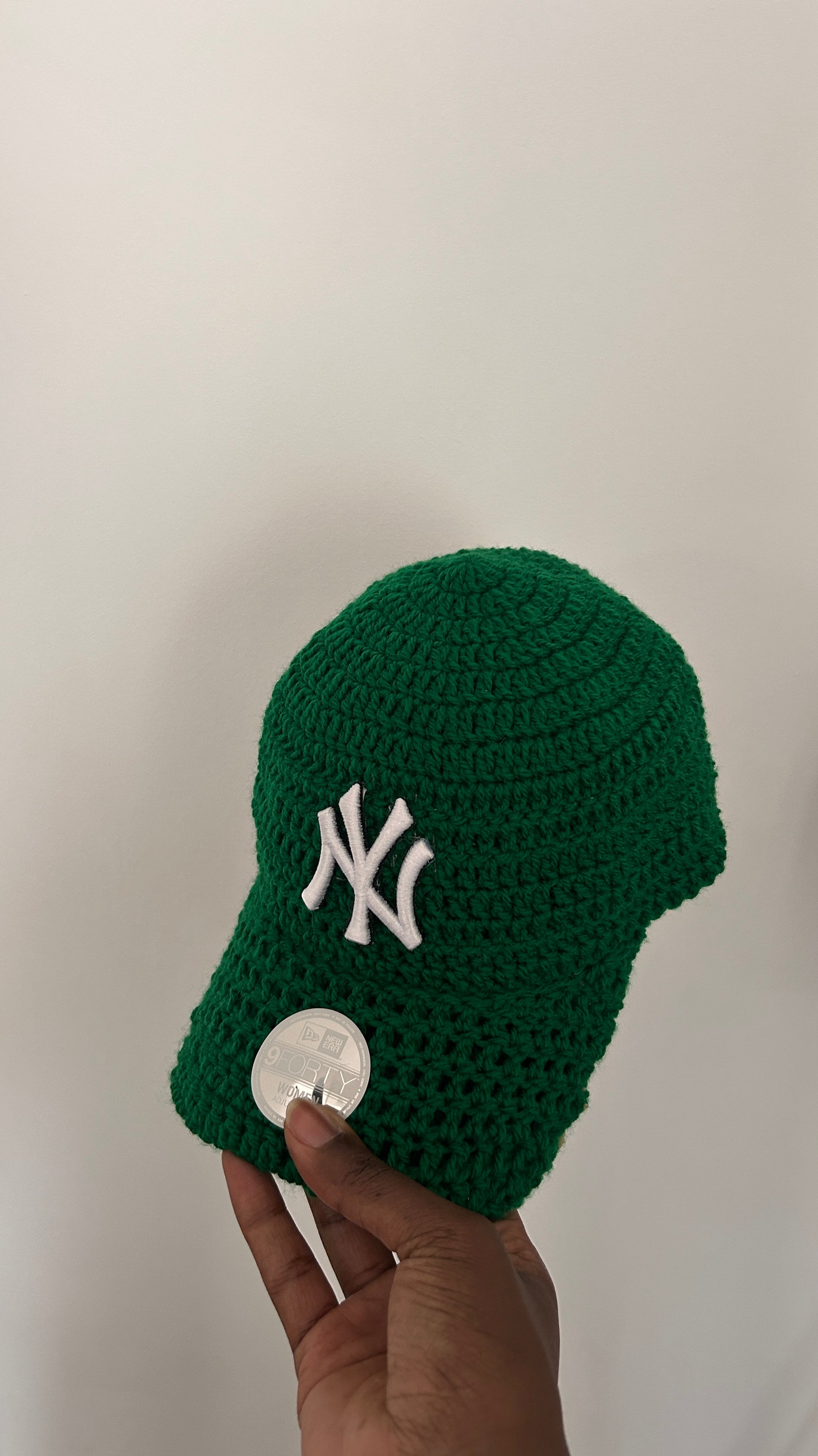 Green knitted baby hat with a white logo held by a hand against a plain background