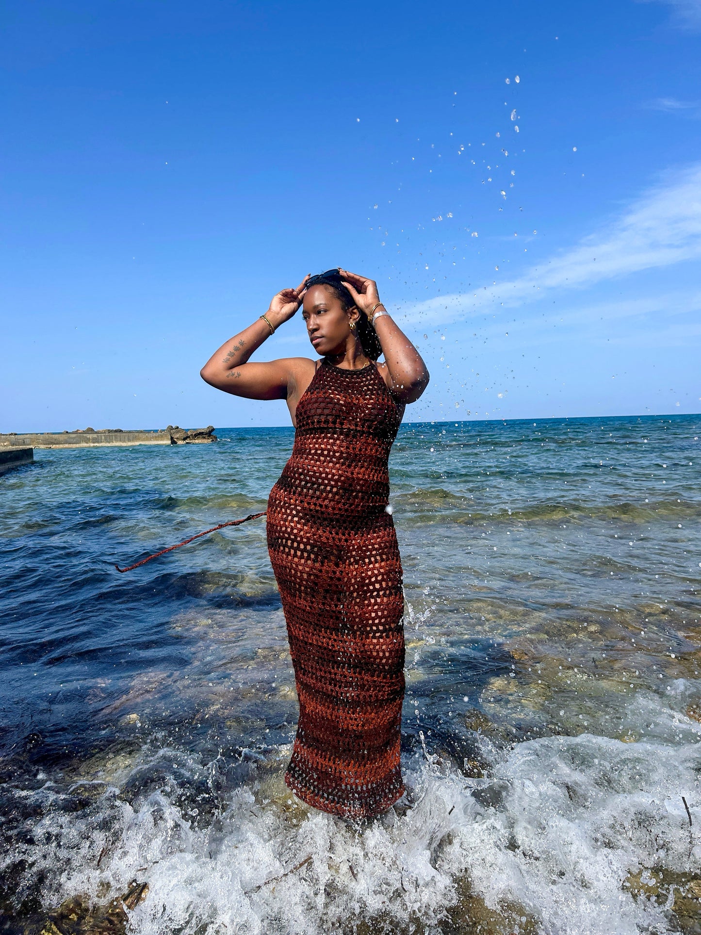 Model wearing a handmade crochet mesh dress standing in shallow sea water, featuring an open-stitch design and full-length silhouette.