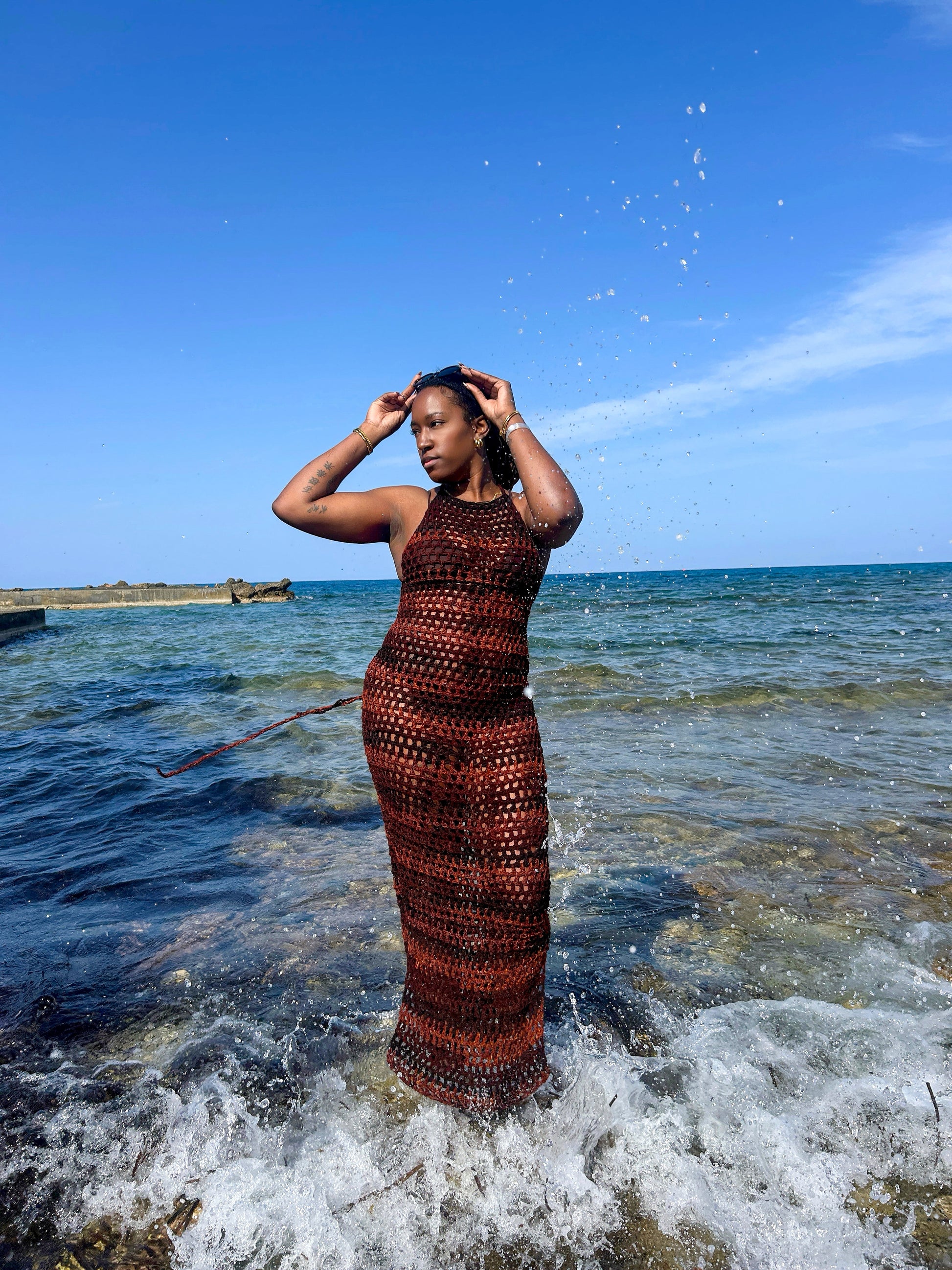 Model wearing a handmade crochet mesh dress standing in shallow sea water, featuring an open-stitch design and full-length silhouette.