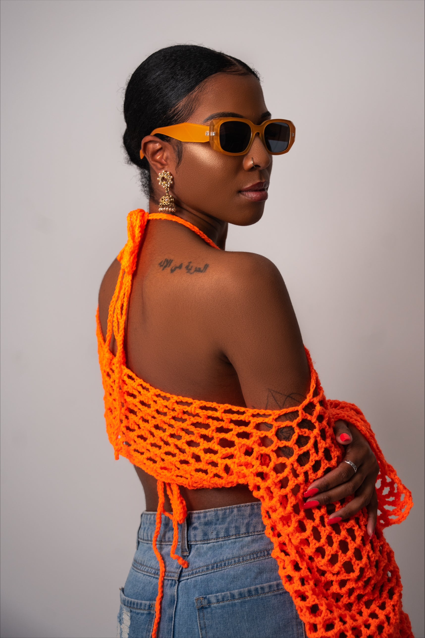 A person wearing an orange crochet shrug and bikini top set, with sunglasses, against a neutral background.