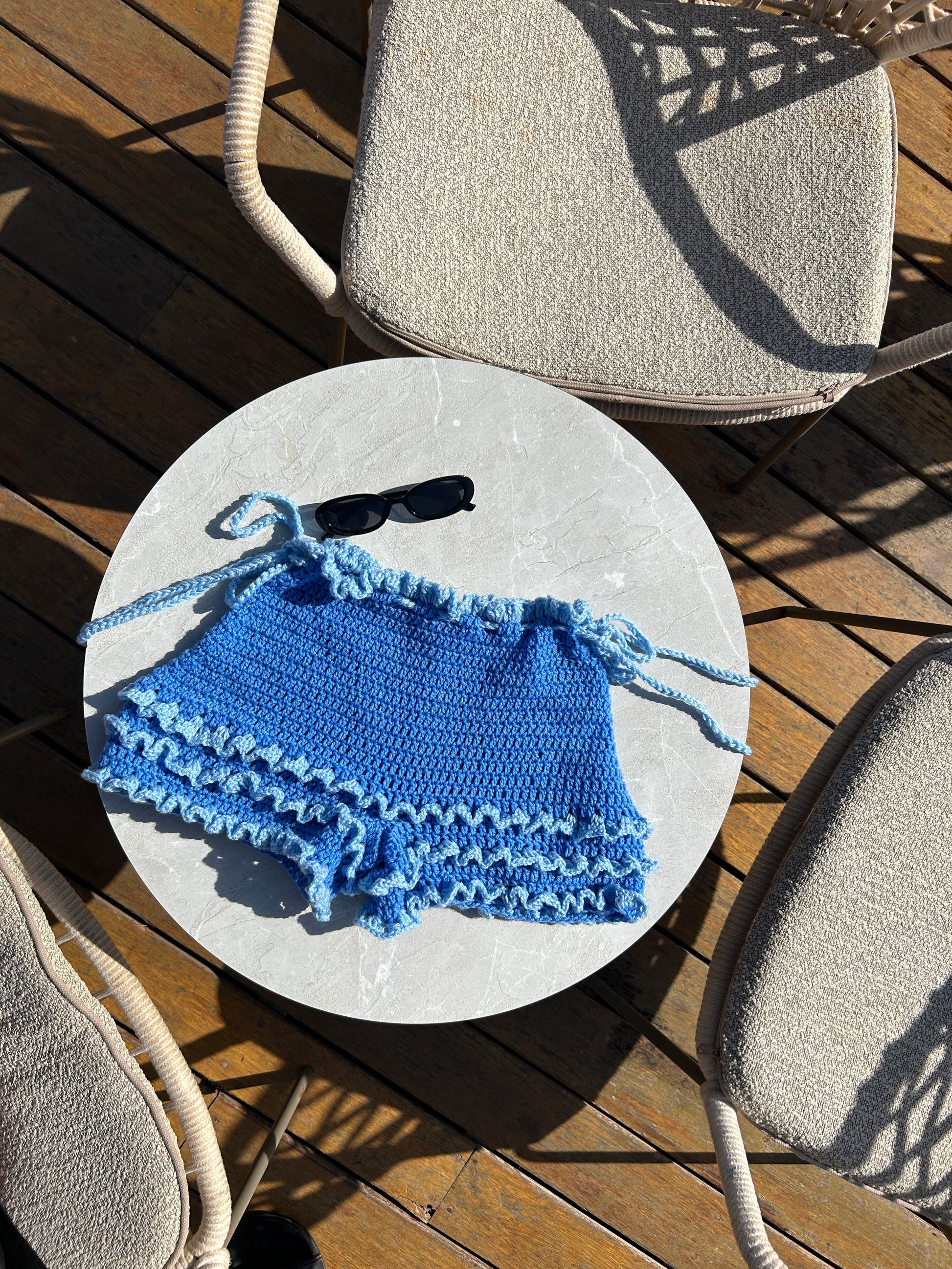Handmade blue crochet shorts with layered ruffle detailing laid flat on a round table outdoors.
