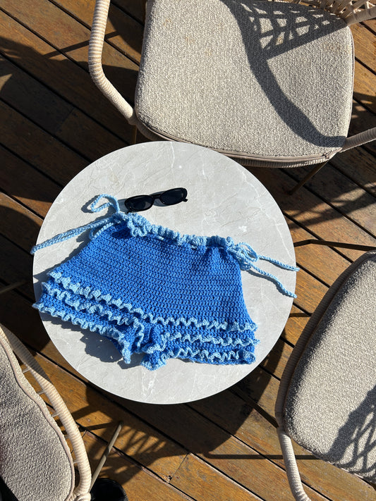 Handmade blue crochet shorts with layered ruffle detailing laid flat on a round table outdoors.