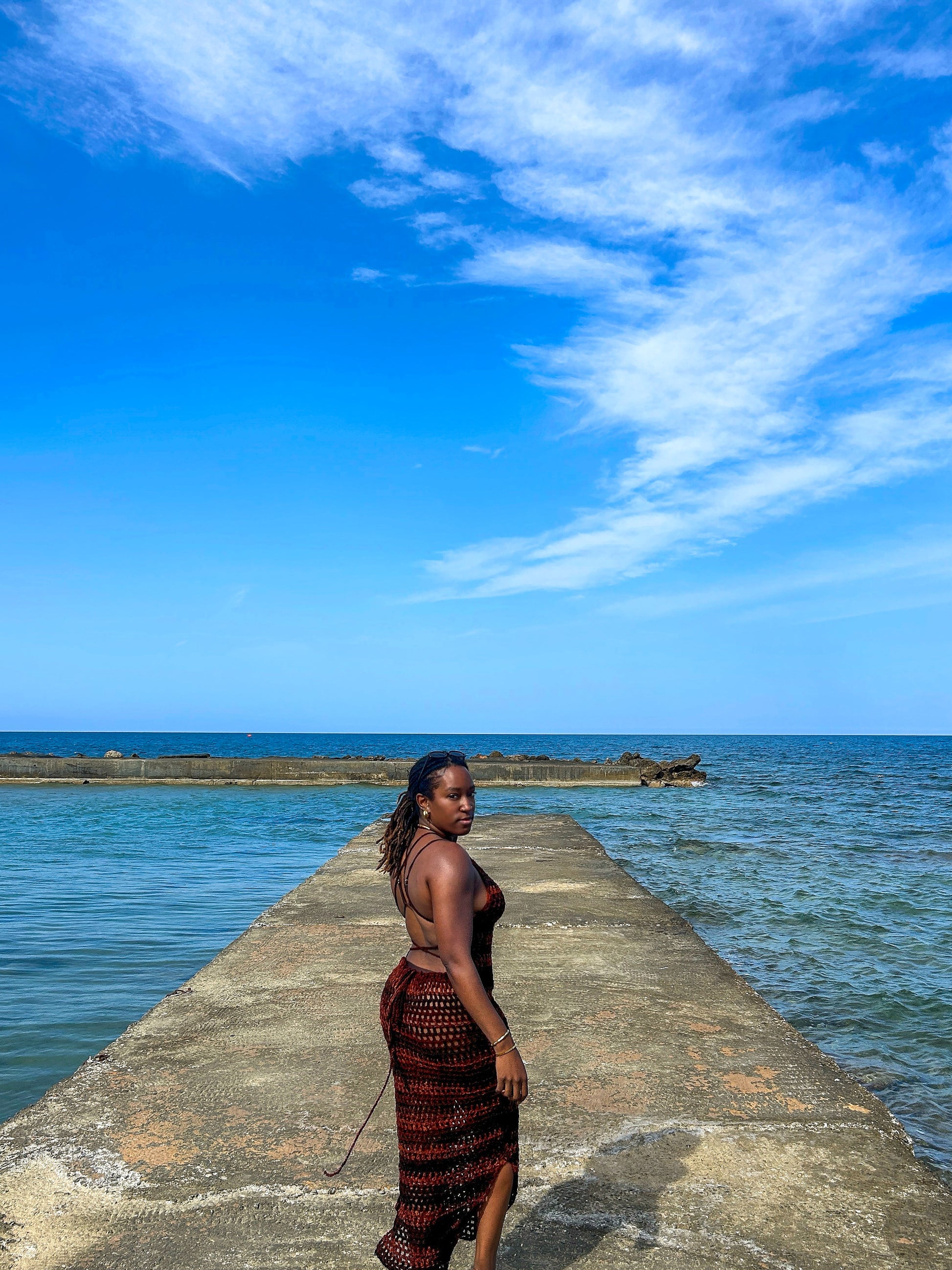 Back view of a handmade crochet mesh dress with open back and tie detailing, photographed on a concrete pier by the sea.

