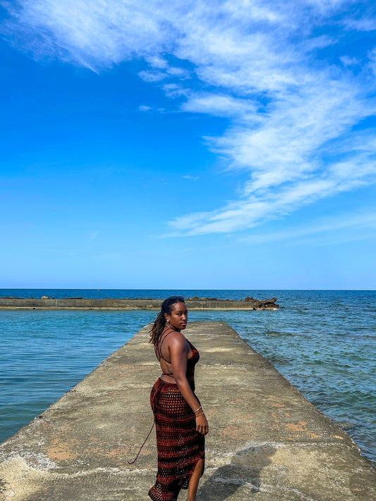 Back view of a handmade crochet mesh dress with open back and tie detailing, photographed on a concrete pier by the sea.
